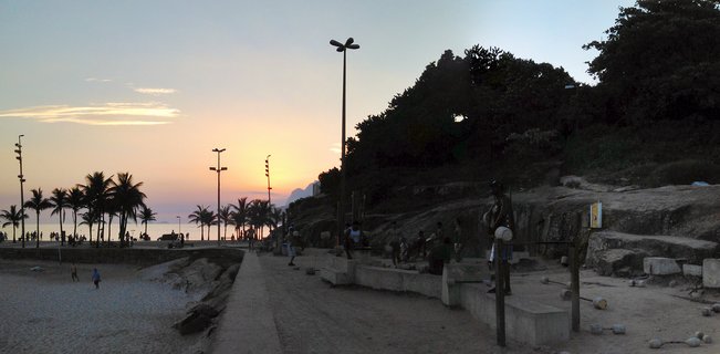 Beach promenade at sunset in Rio de Janeiro with silhouetted palm trees, street lamps, people walking on sand and pathway, rocky hillside with vegetation on the right, colorful orange and pink sky