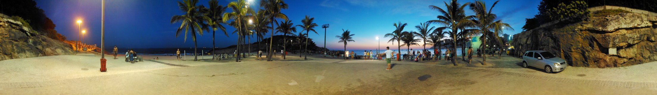 Panoramic beach scene at twilight in Rio de Janeiro showing palm trees, people walking and sitting on sand, illuminated streetlights, rock formations on both sides, and a silver car parked on the right with deep blue evening sky