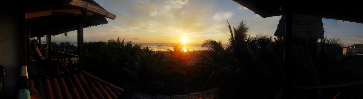 Sunset view over ocean from terrace with silhouetted palm trees and thatched-roof structures on both sides, golden sky with clouds