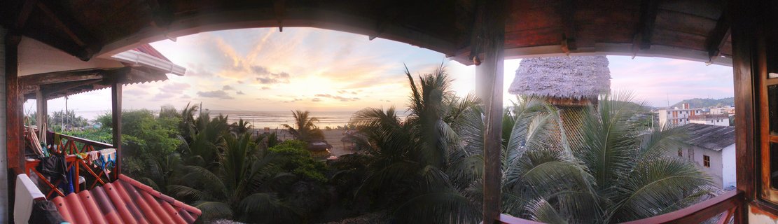 View from wooden balcony overlooking palm trees, tropical buildings with thatched roofs, and Pacific Ocean at sunset in Canoa, Ecuador