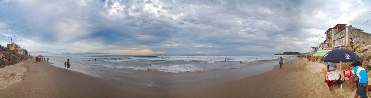 Wide beach scene showing ocean waves, people walking on sand and swimming, beach umbrellas, and buildings along the shoreline at Montanita, Ecuador