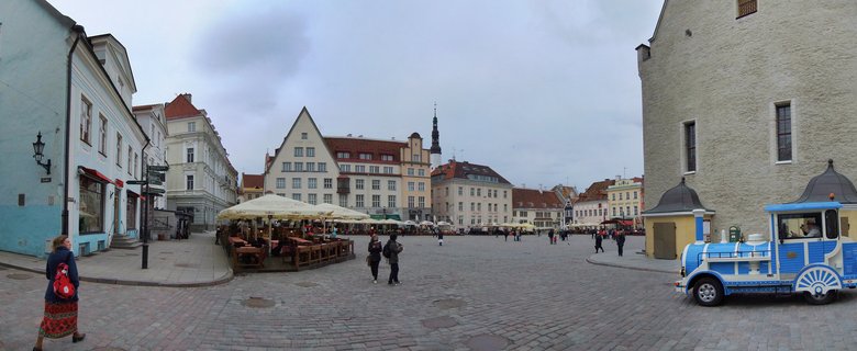 Panoramic view of Tallinn's Town Hall Square showing medieval buildings with red roofs around a cobblestone plaza with outdoor café seating, pedestrians, and a blue tourist train