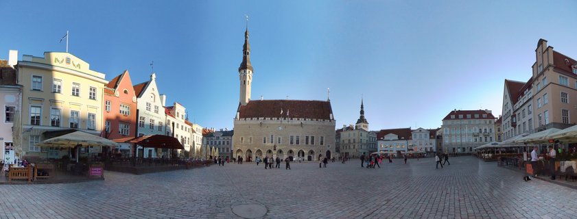 Panoramic view of Tallinn Town Hall Square showing the Gothic Town Hall building with tall spire, colorful medieval houses, cobblestone pavement, and people walking under clear blue sky