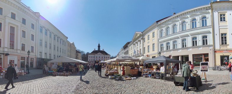 Panoramic view of outdoor market with white tents on cobblestone square surrounded by historic pastel buildings in Tartu Estonia