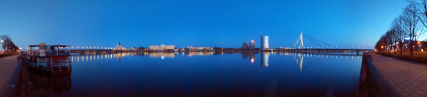 Panoramic view of Riga city skyline at blue hour with illuminated Vanšu cable-stayed bridge over the Daugava River, city lights reflecting on calm water