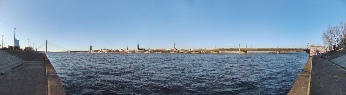 Panoramic cityscape of Riga, Latvia showing the Daugava River with the Stone Bridge and Vanšu Bridge spanning the water. The historic Old Town skyline features multiple church spires including Riga Cathedral and St. Peter's Church against a clear blue sky. Concrete river embankments and bare spring trees line the foreground.