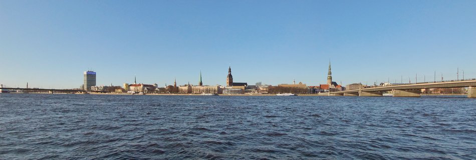 Panoramic cityscape of Riga Old Town viewed from across the Daugava River, showing multiple church spires, historic buildings, bridges, and blue water in the foreground under a clear sky