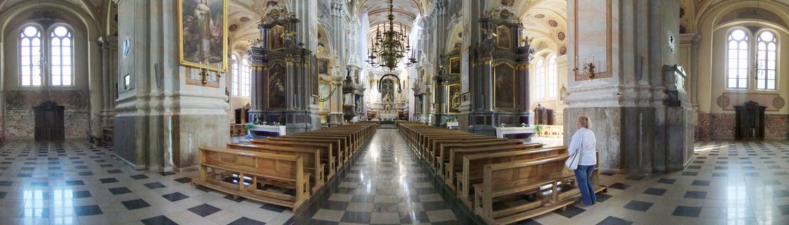 Interior view of a historic cathedral with checkered black and white marble floor, wooden pews, ornate Baroque altars, stone columns, vaulted ceilings with frescoes, and a large chandelier hanging from the center