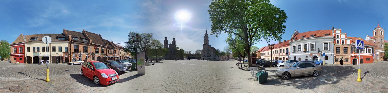Wide panoramic view of a European town square with historic buildings, a cathedral with twin towers, cobblestone ground, parked cars, and trees under a bright blue sky