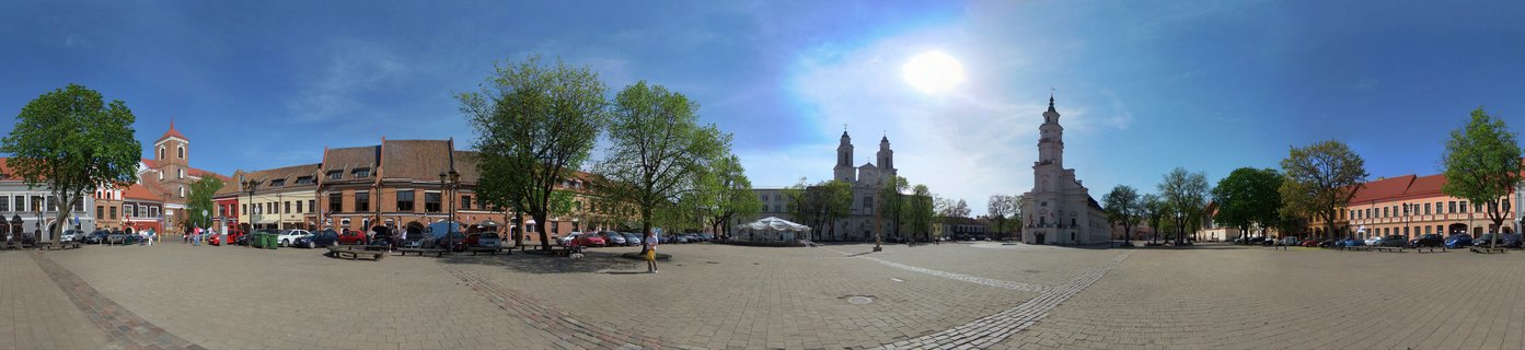 Wide panoramic view of Kaunas Town Hall Square showing historic buildings with red tile roofs, church towers, paved cobblestone plaza, green trees, and bright blue sky with sun