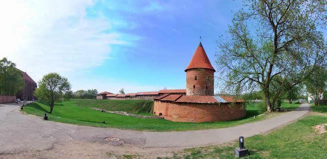 Medieval red brick castle fortress with conical tower, defensive walls, green lawn, trees, and blue sky in Kaunas, Lithuania
