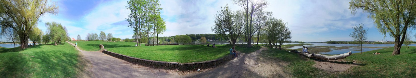 Panoramic view of a riverside park in Kaunas with walking paths, green lawns, trees with spring foliage, and people sitting on grass and logs near the Nemunas River under a partly cloudy sky