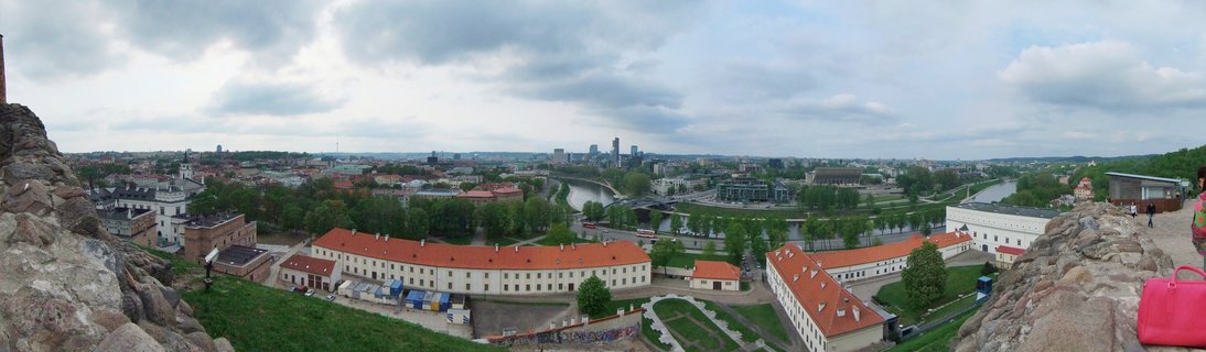Wide panoramic cityscape of Vilnius showing historic buildings with red roofs, the Neris River winding through the city, and modern skyscrapers in the distance under cloudy skies