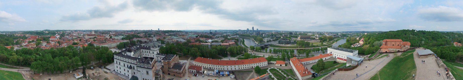 Panoramic cityscape of Vilnius showing historic buildings with red roofs, green trees, the Neris River winding through the city, and modern skyscrapers in the distance under cloudy skies