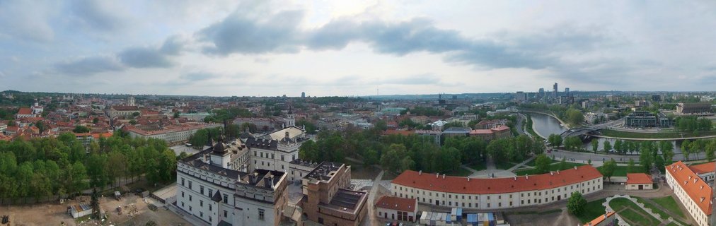 Panoramic aerial view of Vilnius showing historic buildings with red roofs, green trees, the Neris River, and modern skyscrapers in the distance under cloudy sky