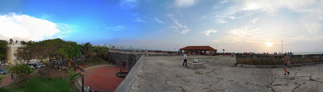Panoramic view of people walking on historic stone fortress walls in Cartagena with Caribbean Sea, colonial buildings, and city skyline at sunset
