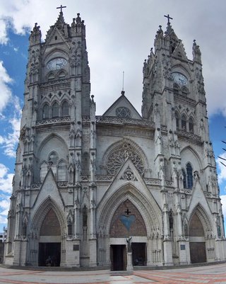 White stone neo-Gothic cathedral with two tall clock towers topped with crosses, featuring pointed arches, rose windows, and ornate facade details against blue sky with clouds