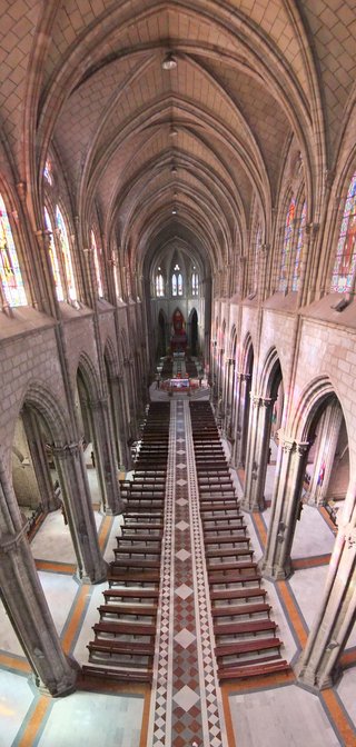 Interior view of a Gothic cathedral showing a long nave with wooden pews, stone columns, vaulted ceiling, stained glass windows, and patterned tile floor leading to an altar