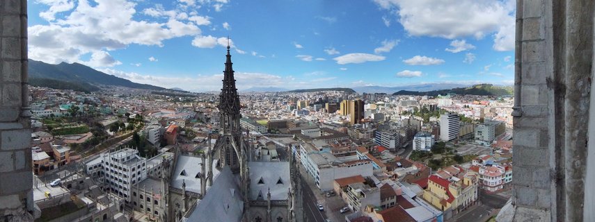 Ornate Gothic church spire in foreground with dense city buildings and mountains in background under blue sky with white clouds