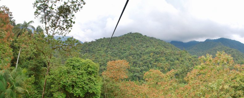 Aerial view of dense green tropical forest covering mountain slopes with cloudy overcast sky above and a cable visible in the upper portion