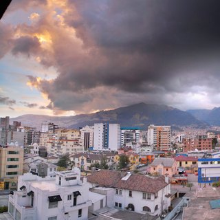 Aerial view of Quito city buildings with terracotta roofs and modern high-rises under dramatic storm clouds with mountains in background at sunset