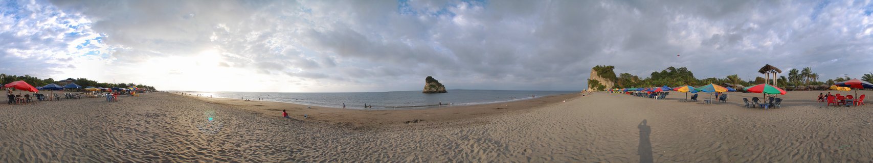 Wide panoramic beach scene showing sandy shore with colorful umbrellas, people walking and swimming, rock formations in the ocean, palm trees, and cloudy sky over Tumaco, Colombia