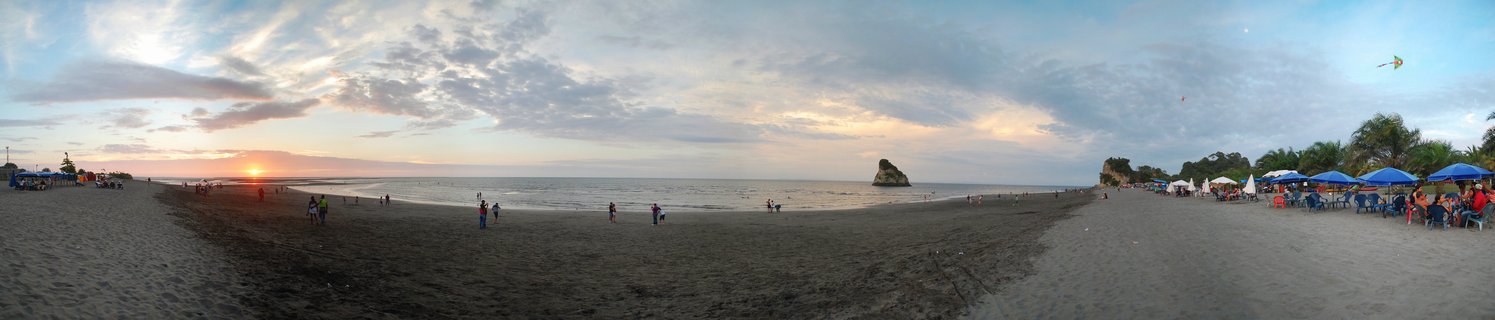 Wide beach scene at sunset showing dark sand, ocean waves, scattered people, blue vendor tents, palm trees, and a rock formation in the water under a cloudy sky with golden light