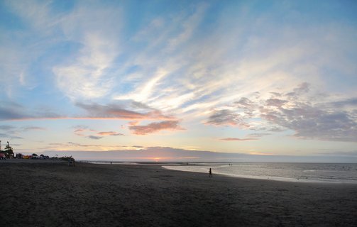 Wide beach panorama at sunset showing ocean, dramatic cloudy sky with pink and orange tones, dark sand, and small silhouettes of people walking along the shoreline in Tumaco, Colombia