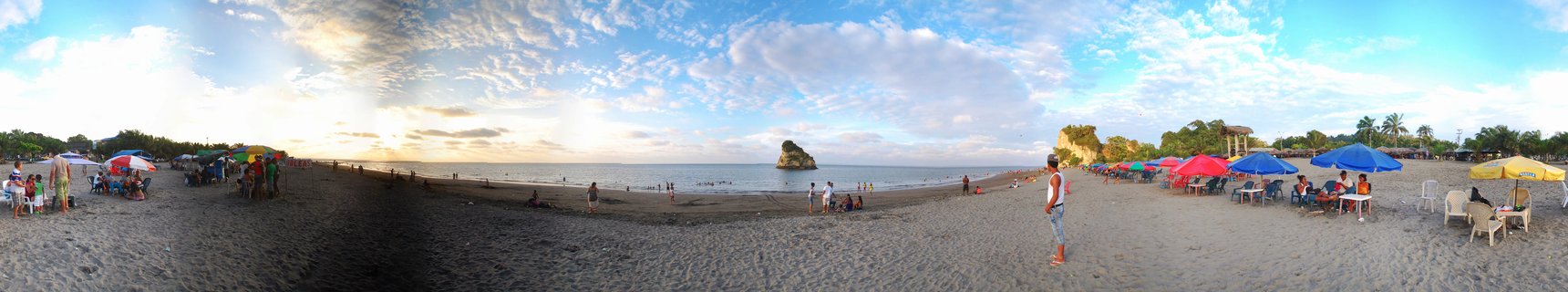 Wide beach panorama showing people under colorful umbrellas on dark sand with ocean, rock formation, and cloudy sky at Tumaco, Colombia