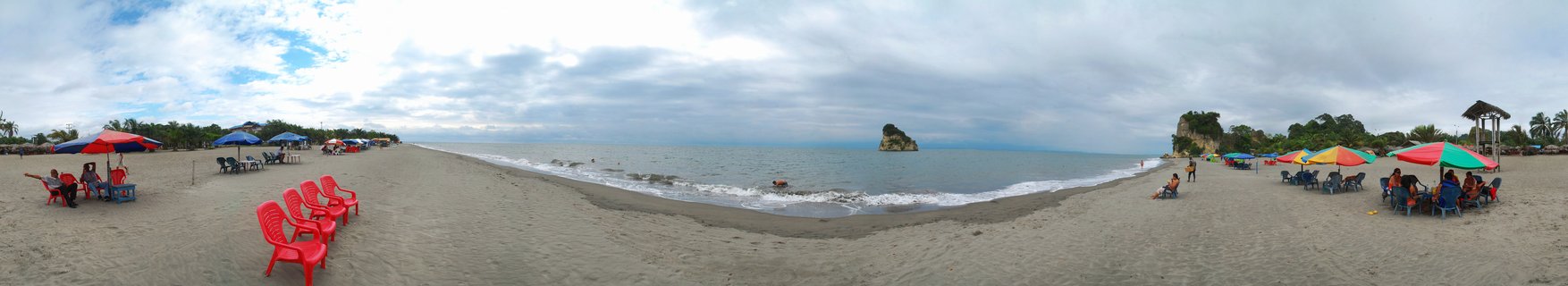 Wide panoramic beach scene showing colorful umbrellas and red chairs on dark sand, people relaxing by the ocean, with a rock formation visible in the water under cloudy sky