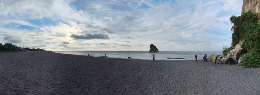 Wide beach scene showing dark volcanic sand stretching to calm ocean waters with a large rock formation in the distance, several people walking along the shore, vegetation-covered cliff on the right side, and cloudy sky above