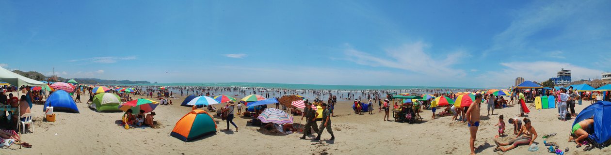 Wide panoramic view of a crowded beach with numerous colorful umbrellas and tents, people swimming in the ocean, and buildings visible along the coastline under a clear blue sky.