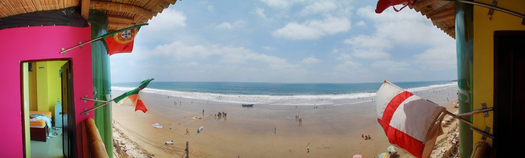 Panoramic beach scene viewed between colorful pink and yellow building walls with flags, showing sandy beach, ocean waves, and scattered people under blue sky with clouds