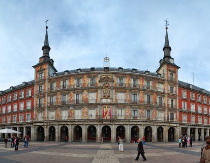 Wide view of Casa de la Panadería building at Plaza Mayor in Madrid with red painted facade, decorative frescoes, two corner towers with spires, stone arches at ground level, and people walking on cobblestone plaza