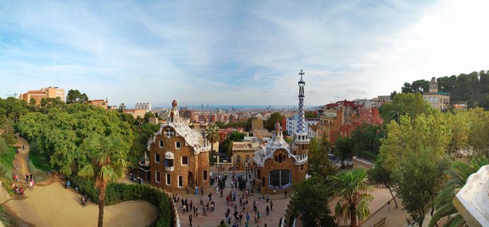 Panoramic view of Park Güell's entrance featuring two ornate mosaic gatehouses with undulating white stone roofs, a tall blue and white checkered spire tower, crowds of tourists on the plaza below, lush green trees and palm trees surrounding the area, with Barcelona city buildings and the Mediterranean Sea visible on the horizon under a partly cloudy blue sky during late afternoon