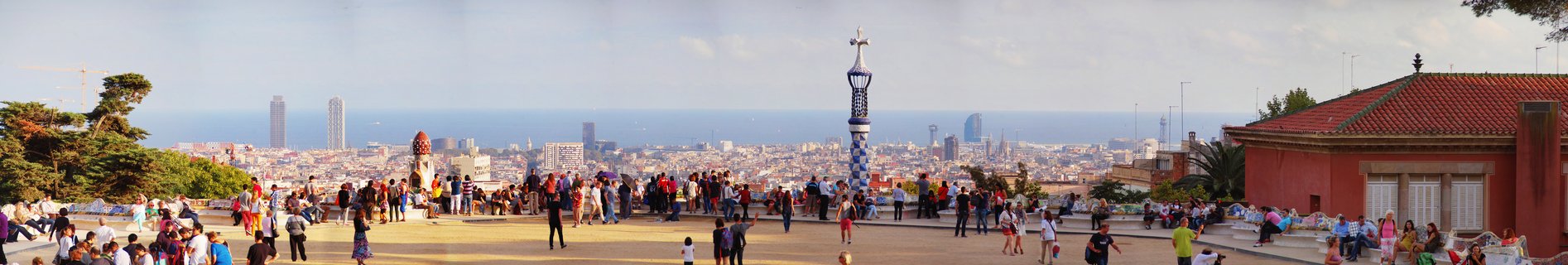 Wide panoramic view from Park Güell terrace showing crowds of tourists, Gaudí's distinctive towers with crosses, red-tiled buildings, and Barcelona cityscape extending to the Mediterranean Sea