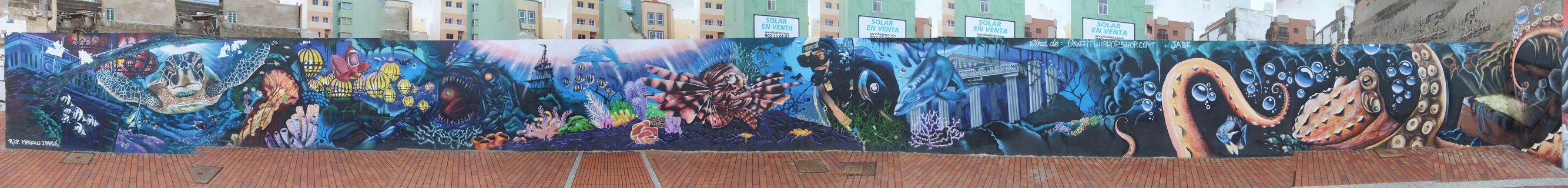 Panoramic street art mural showing colorful underwater scene with sea turtles, octopus, lionfish and tropical fish painted on wall along beachfront promenade with apartment buildings behind