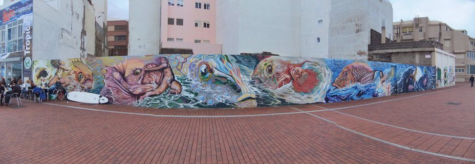 Panoramic photograph of a long colorful mural wall featuring painted fish, ocean waves, and marine-themed street art along a brick-paved waterfront promenade with buildings and outdoor cafe seating visible