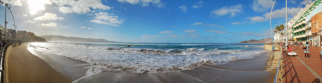 Panoramic beach scene showing Las Canteras promenade with colorful buildings, people walking, golden sand beach, ocean waves, and blue sky with clouds in Gran Canaria, Spain