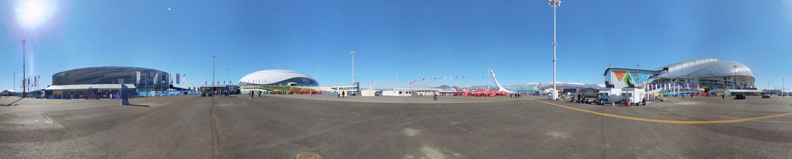 Wide panoramic photograph showing multiple modern sports stadium buildings in Sochi Olympic Park under bright blue sky with paved plaza in foreground