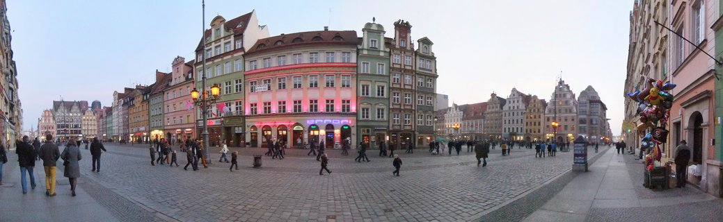 Panoramic view of Wrocław Market Square at dusk showing colorful historic buildings, cobblestone ground, and people walking across the square with street lamps illuminated