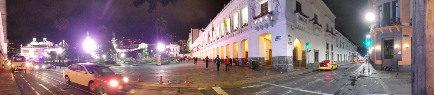 Night panoramic view of a colonial plaza with illuminated white government building, pedestrians walking, cars on wet streets, and historic architecture in Quito, Ecuador