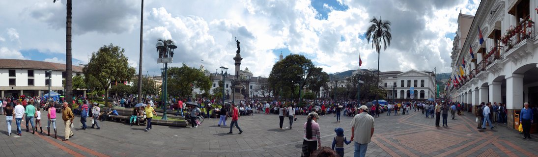 Wide panoramic view of a bustling colonial plaza with a central independence monument, white colonial buildings with arcades, palm trees, and numerous pedestrians under a cloudy sky