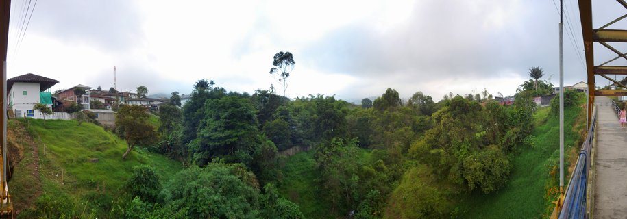 Wide panoramic photograph showing a green valley in Salento Colombia with houses on hillsides, dense trees and vegetation, and a yellow suspension bridge on the right side with a person walking across under cloudy skies