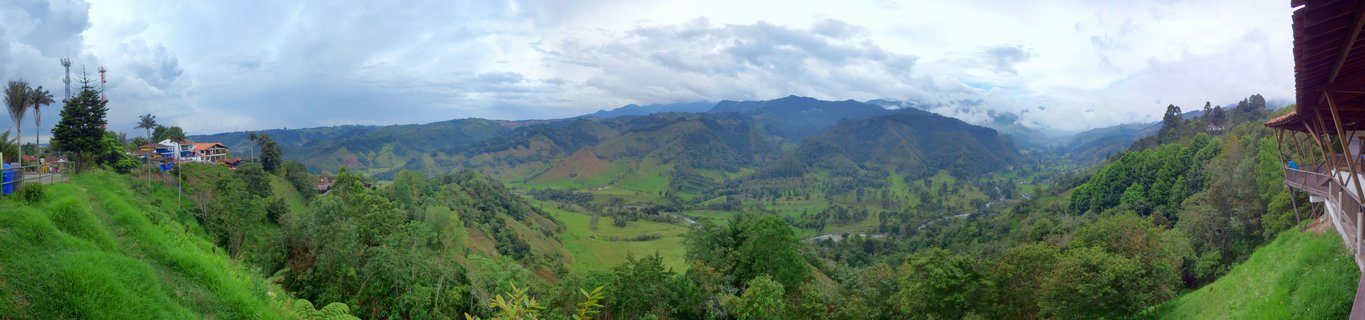 Wide panoramic landscape showing green mountain valley with coffee plantations, scattered buildings with red roofs, dense tropical vegetation, and cloudy sky in Salento, Colombia