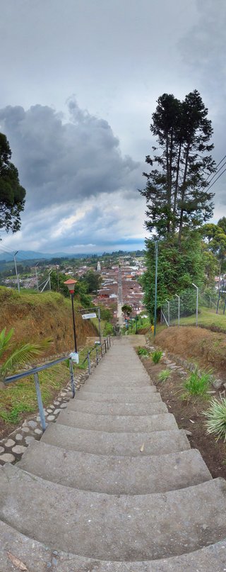 Long concrete staircase with metal railings descending toward a colonial Colombian town with red-tile roofs, church tower, and green mountains under cloudy sky