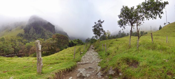 A stone path leads through green rolling hills with wooden fence posts, trees scattered across the landscape, and fog covering mountain peaks in the background