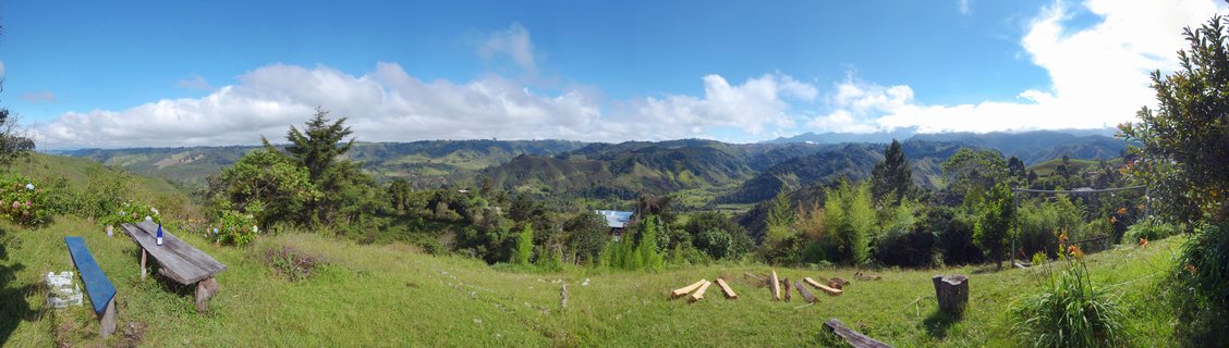 Wide panoramic landscape showing green rolling mountains and hills in Colombia's coffee region near Pereira, with wooden picnic benches in the grassy foreground under a blue sky with white clouds