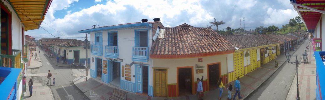 Panoramic street view of colonial buildings with blue, yellow, and white facades, terracotta roofs, people walking on sidewalks, cloudy sky above