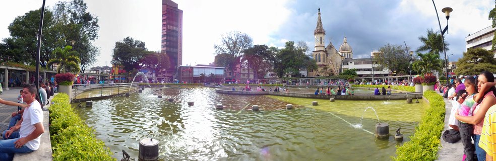 Wide panoramic view of a public plaza with a large ornamental fountain, people sitting along the edges, a church with tall spire in the background, and urban buildings surrounding the square